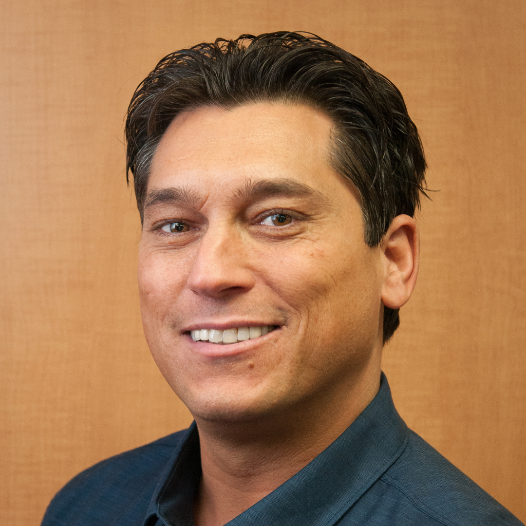 Employee headshot of Nick Mangiardi with a blue shirt and brown background. 