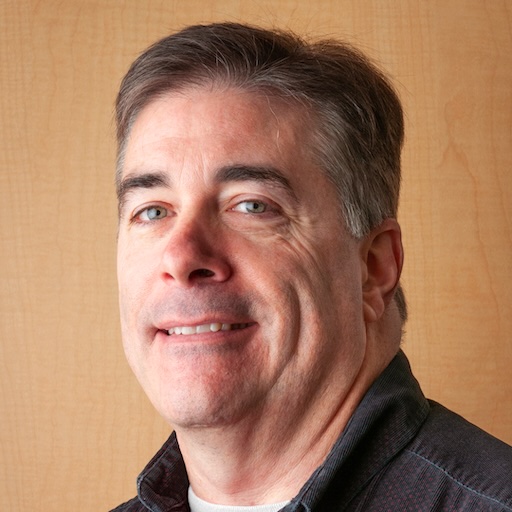 A headshot photo of John Byrne from standing against a light brown background.